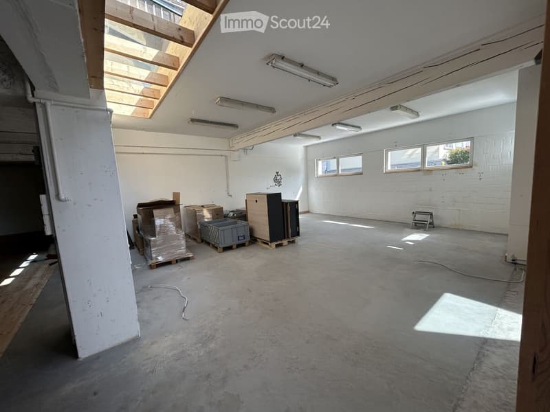 Empty room with concrete floor, white walls, exposed wooden beams, and skylight. Some storage boxes and a ladder are present.