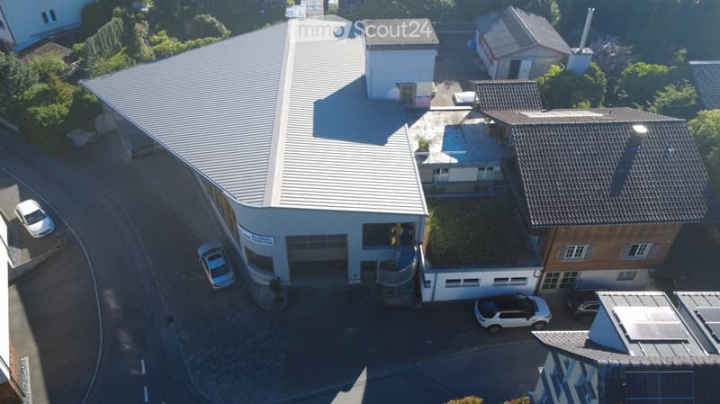 Aerial view of a modern industrial building with a large garage, surrounded by several parked cars and neighboring structures.