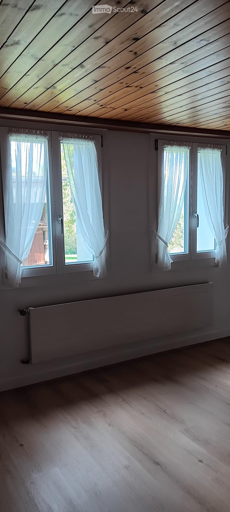 white walls, wooden ceiling, three windows with white curtains, a white radiator, wooden floor