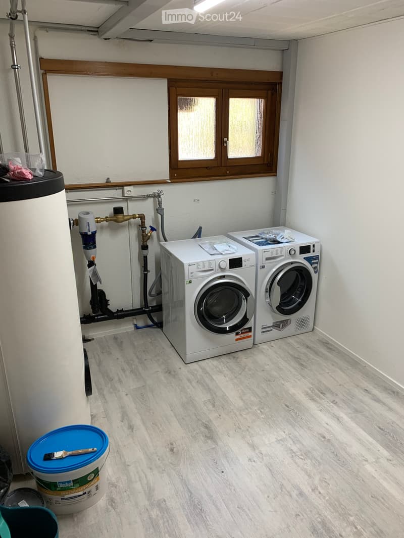 Bathroom with wooden flooring, white walls, and a window. Contains a washing machine, a drum dryer, a water heater, and a bucket with a paintbrush on top.