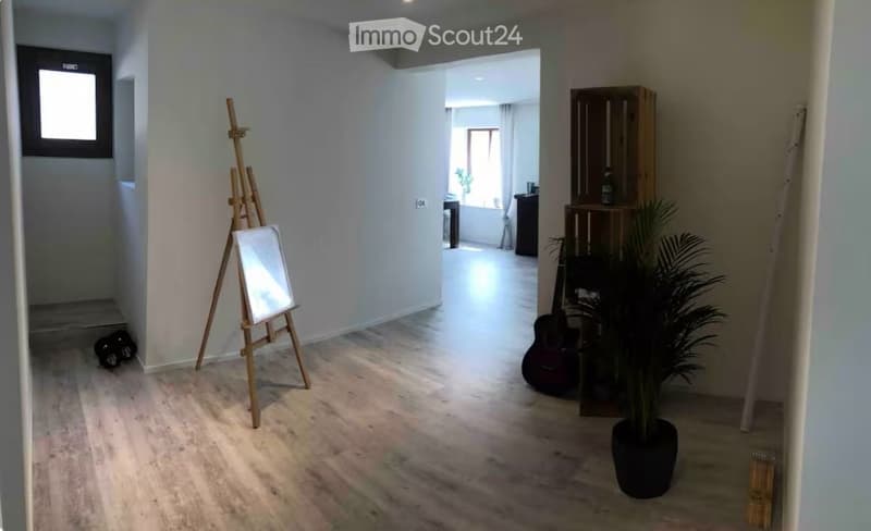 The room features a wooden floor, white walls, and a large window. Visible objects include an easel with a canvas, a guitar, a potted plant, and a wooden cabinet.