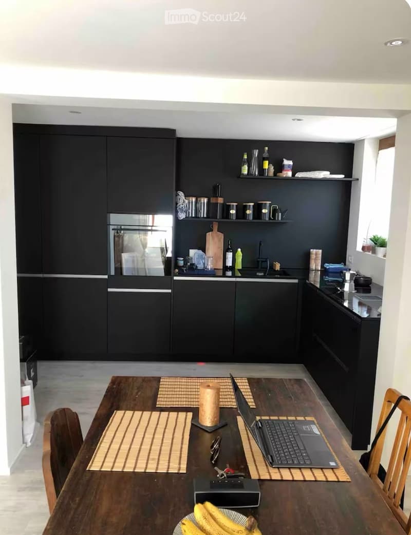 Modern kitchen with black cabinets, stainless steel appliances, and a wooden dining table with chairs. The table has a laptop, bananas, and other small items.