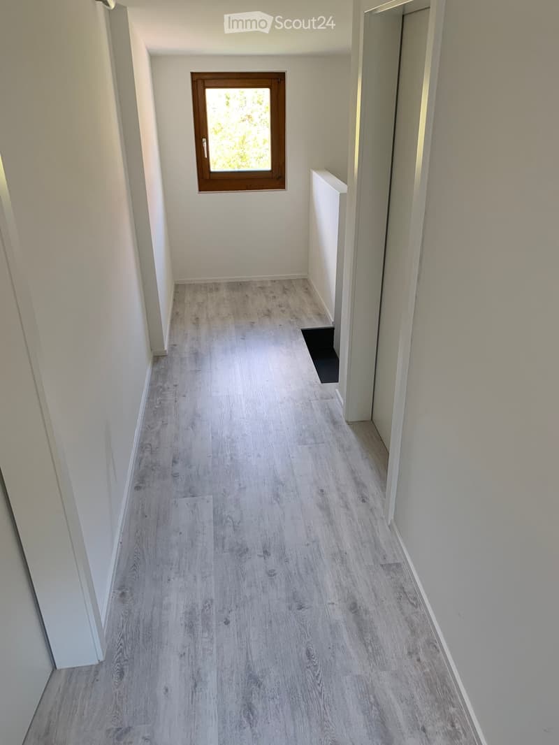 Empty hallway with light grey wooden flooring and white walls, featuring a single window with a wooden frame on one wall and an open doorway on the opposite side.