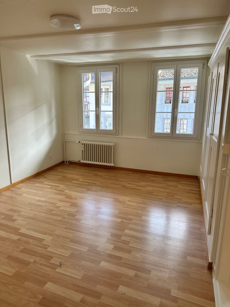 Empty room with light wooden flooring, white walls, and large windows with grey frames. A radiator is visible against one wall.