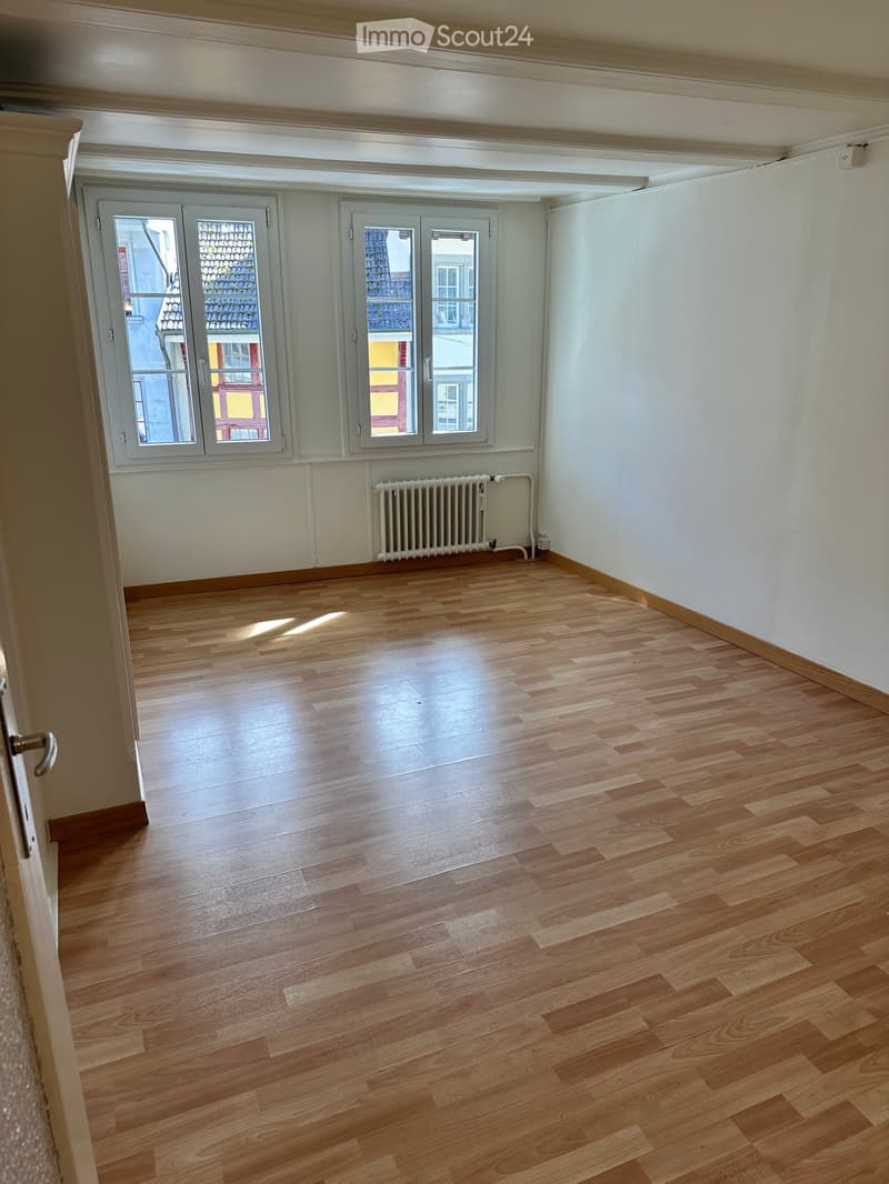Empty room with wooden flooring, white walls, and large windows with grey frames. A radiator is visible against one wall.