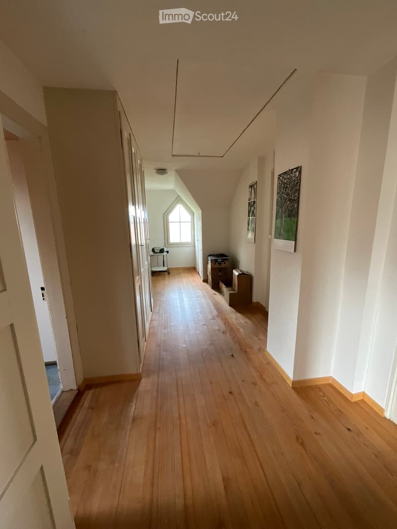 A narrow hallway with wooden flooring, white walls, and a slanted ceiling. There are doors on the left and a window at the end, with some stacked boxes and a small cart visible.