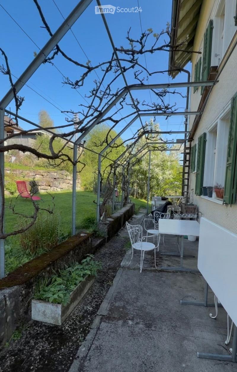 Balcony with metal frame, white table and chairs, potted plants, and a view of a garden and stone wall.