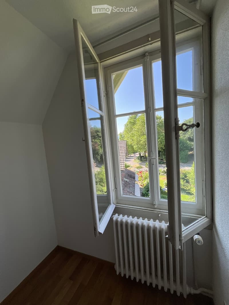 Empty room with wooden flooring, white radiator, and large windows offering a view of a garden and trees.