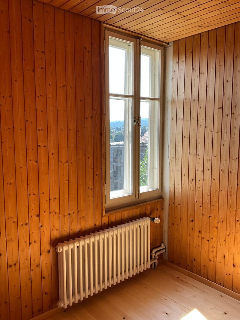 Empty room with wooden walls and floor, featuring a radiator and windows with a view of the outside.