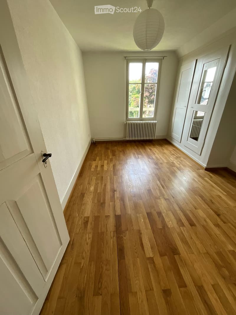 Empty room with wooden flooring, white walls, and large windows letting in natural light. Features a radiator and a hanging light fixture.