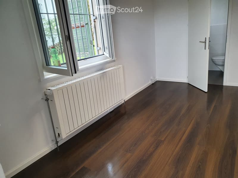 empty room, white walls, white radiator, dark wooden floor, window with grill, toilet in adjacent room