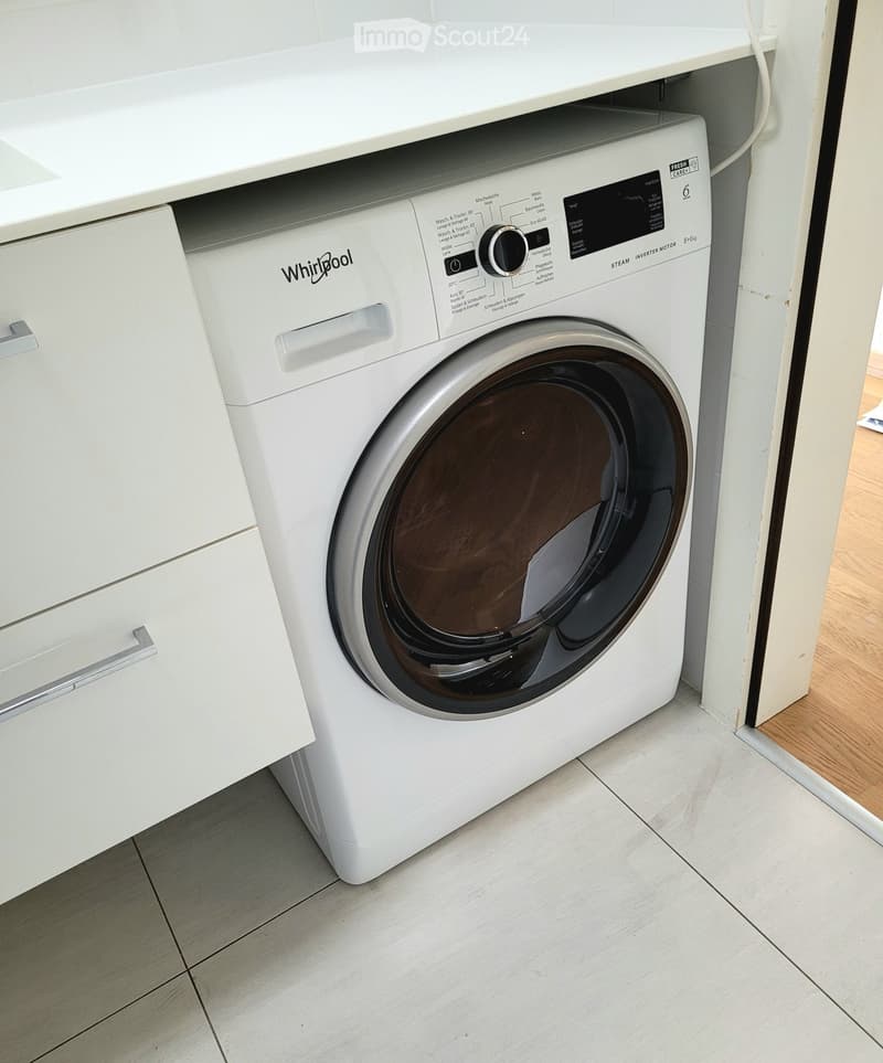 Bathroom. A white Whirlpool washing machine is installed in a corner of a room with light-colored tiled flooring and white cabinetry.