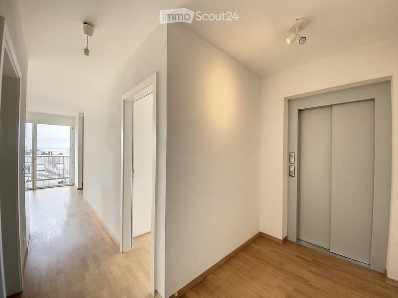 Corridor / Hallway. Empty hallway with wooden flooring, white walls, and recessed lighting. Features an elevator door on the right and a sliding glass door leading to a balcony on the left.