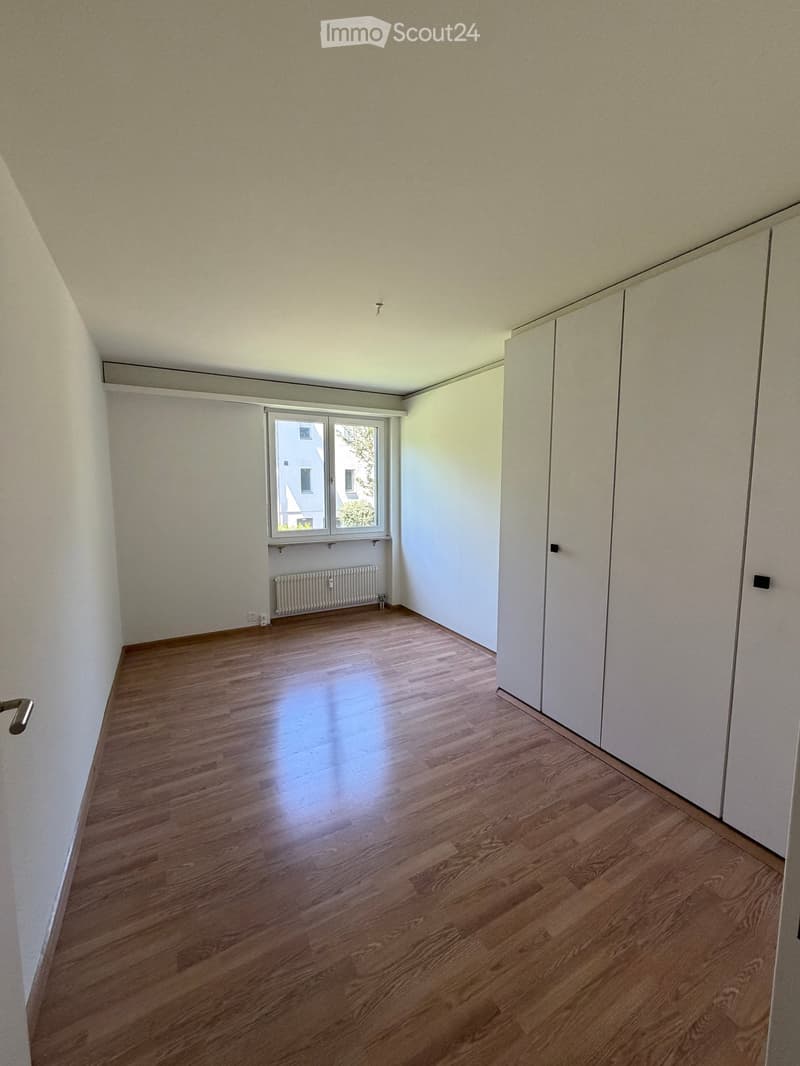 Empty room with wooden flooring, white walls, and a window with a radiator below it. The room has built-in white cabinets on one side.