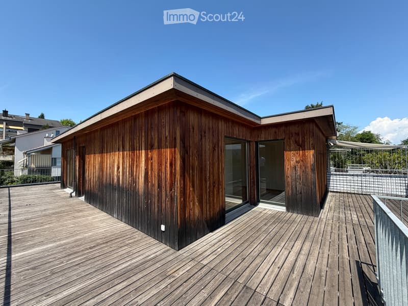 Modern wooden structure with large glass windows and a wooden deck, surrounded by greenery and a clear blue sky.