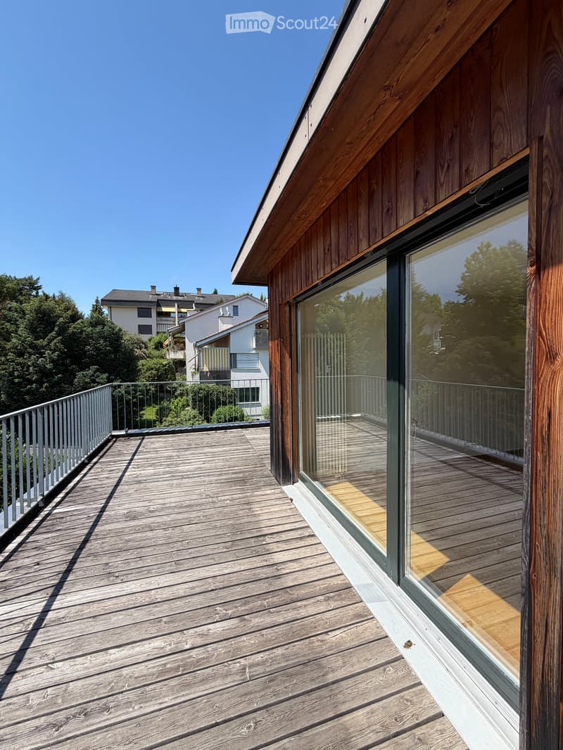 Wooden deck with glass railing, overlooking a residential area with trees and neighboring houses.