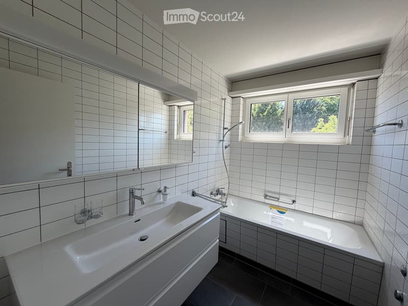 Modern bathroom with white tiled walls and floor, featuring a large sink, bathtub, and a window with a view of greenery.