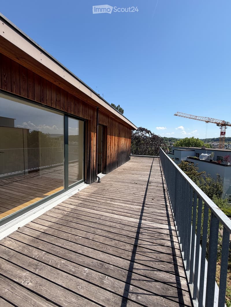 Wooden deck with metal railing, adjacent to a modern building with wooden siding and large glass windows.