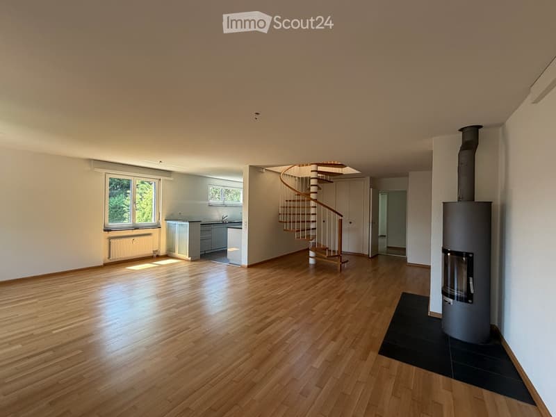 Bright, open-plan space with wooden flooring, a spiral staircase, a kitchen area, and a wood-burning stove. The room features large windows and neutral-colored walls.