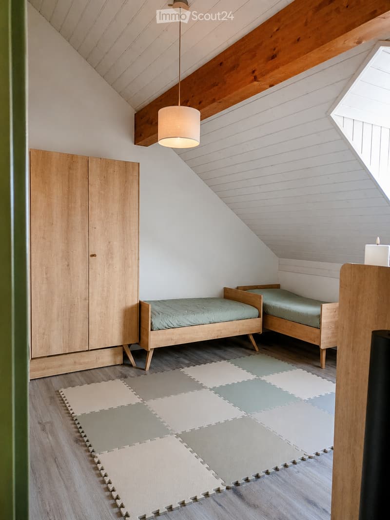 Cozy attic bedroom with two wooden twin beds, a wardrobe, and a hanging lamp. The room features wooden paneling and a slanted ceiling with exposed beams.