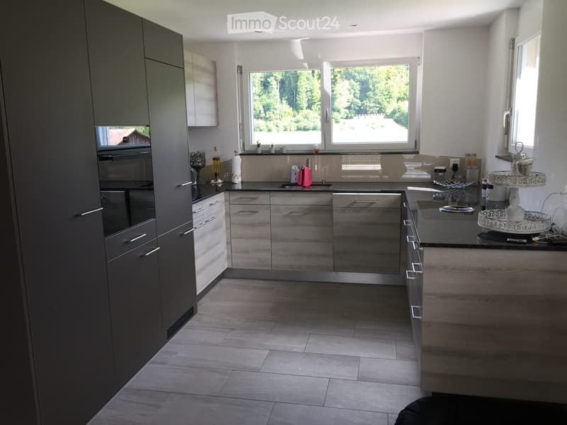 Modern kitchen with grey cabinets, wooden countertops, and large windows overlooking a green area.