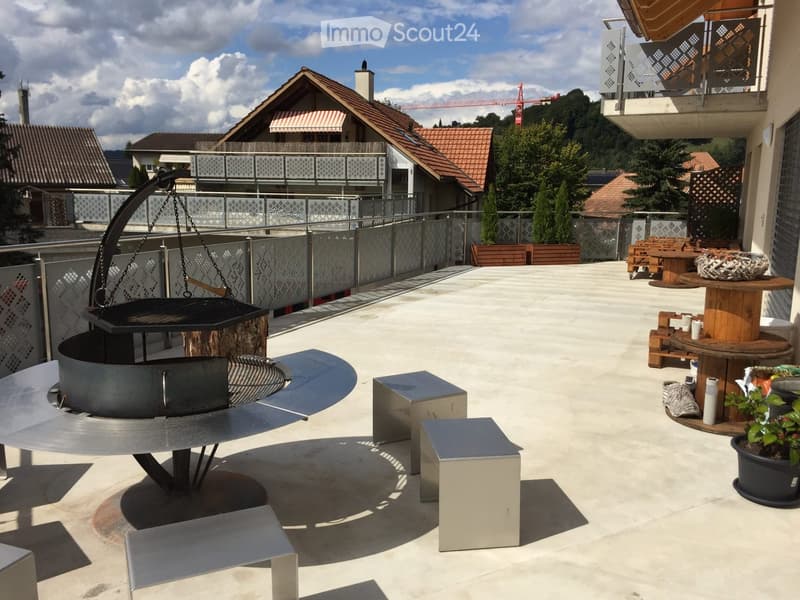 Balcony with a modern metal fire pit, wooden table, and seating area, surrounded by a metal railing and potted plants.