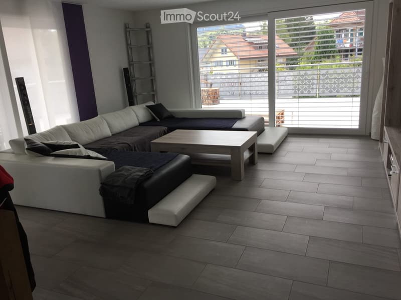Modern living room with grey sectional sofa, coffee table, ottoman, large windows, and balcony access.