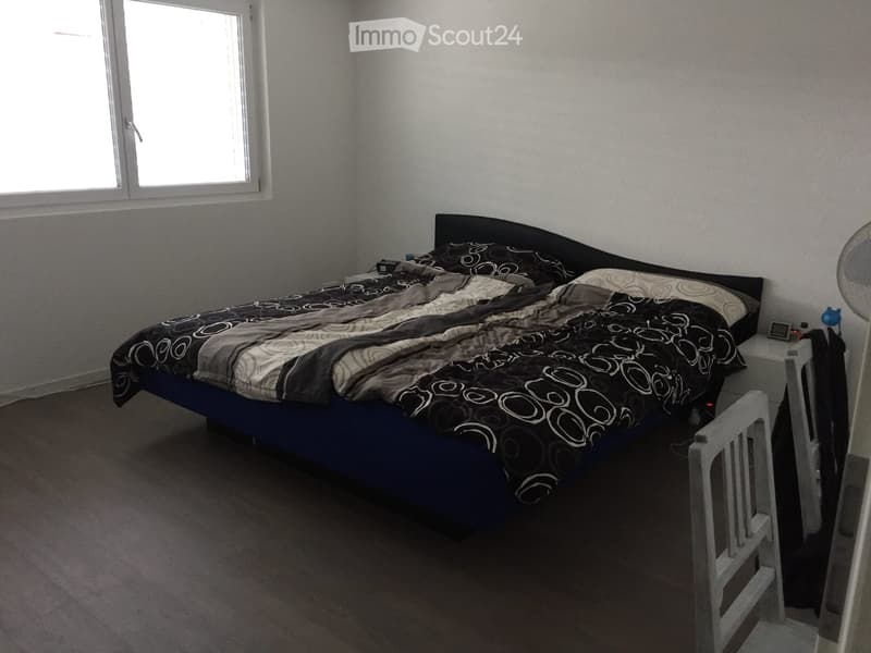 Bedroom with a large bed, nightstand, and fan. The bed has a dark blue frame and a patterned duvet. The room has wooden flooring and a window with blinds.