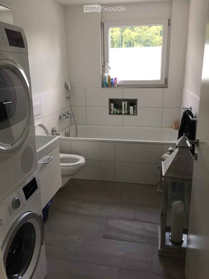 Modern bathroom with a bathtub, toilet, shower, washing machine, and a window with toiletries on the sill.