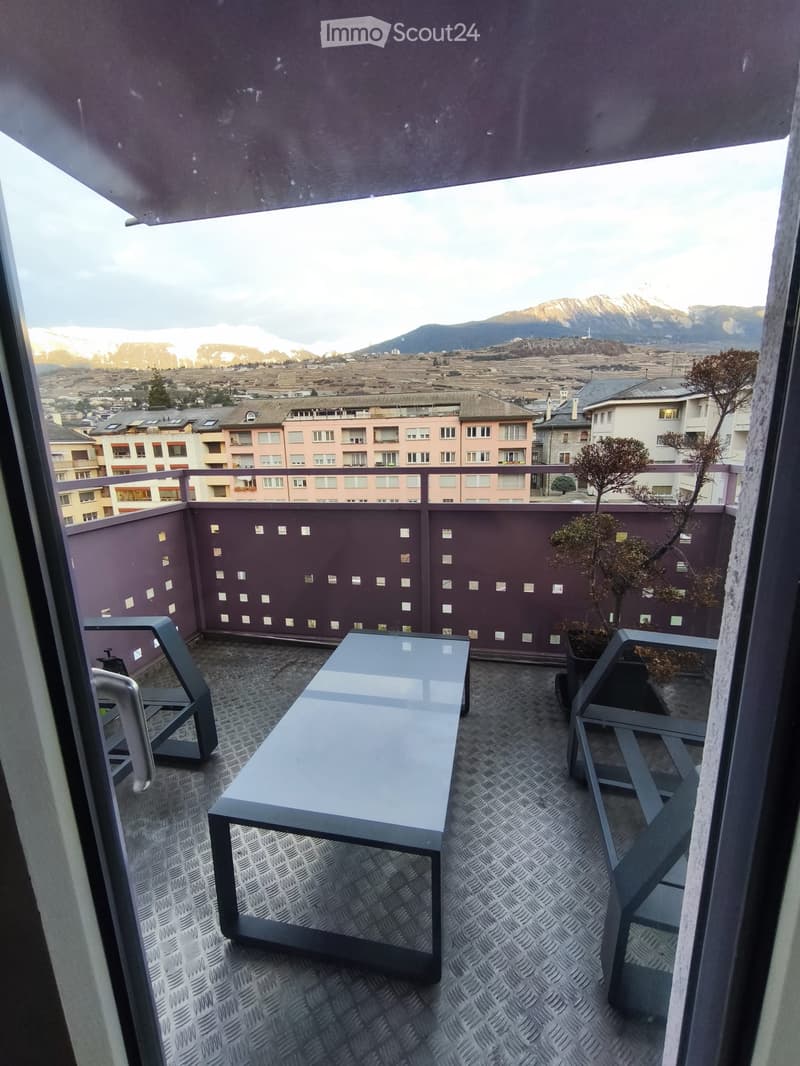Balcony with a modern table and chairs, overlooking a cityscape with mountains in the background.