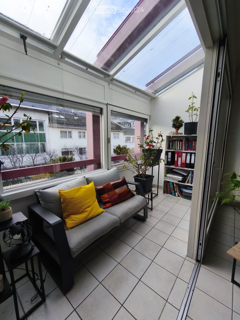 Bright, modern balcony with a grey sofa, colorful cushions, a small table, a bookshelf, and potted plants.