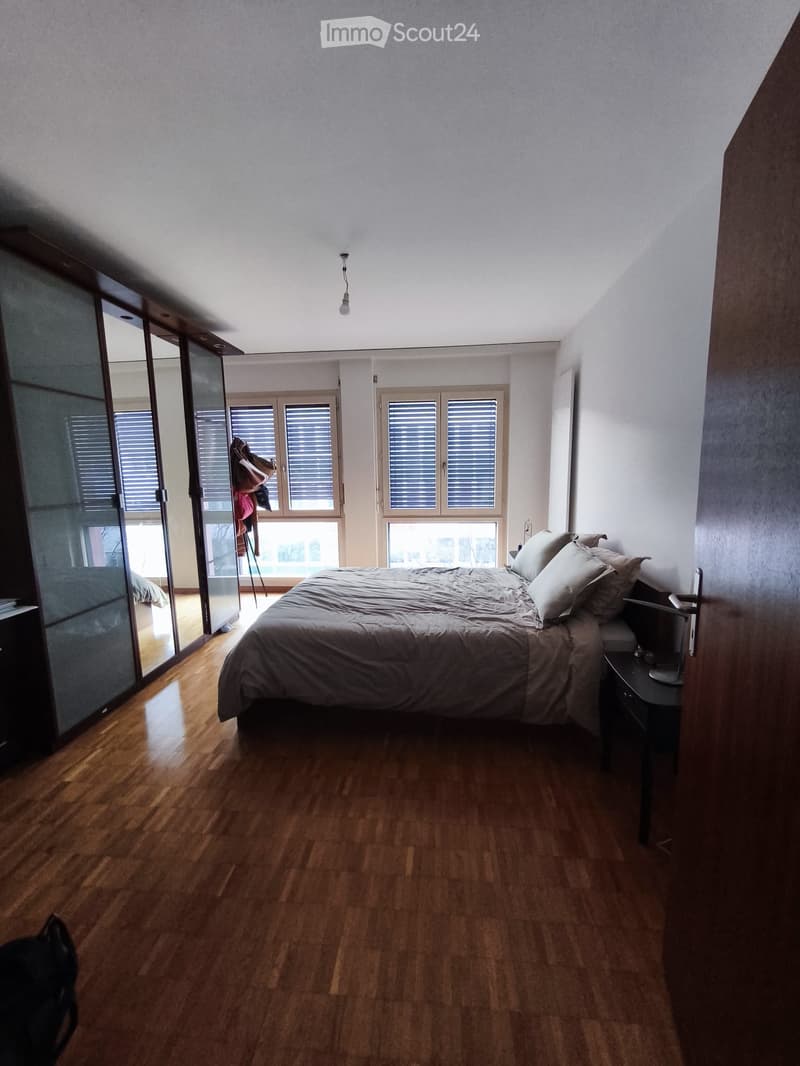 Modern bedroom with wooden flooring, large bed with pillows and duvet, nightstand with lamp, and a mirrored wardrobe with clothes visible inside.
