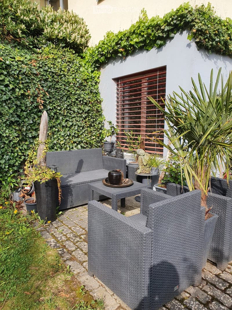 A cozy outdoor seating area with a grey wicker sofa, matching chairs, and a coffee table, surrounded by potted plants and greenery.