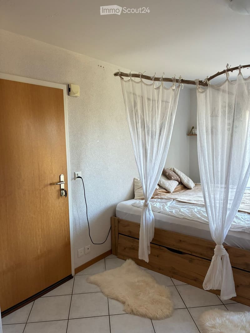 Cozy bedroom with a wooden bed, white bedding, pillows, and a fluffy white rug on the floor. A sheer white curtain is draped over the bed, and a wooden door is visible on the left side.