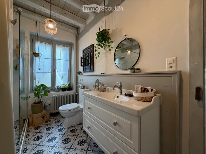 Cozy bathroom with a double sink, mirror, toilet, wicker basket, potted plants, and a hanging light fixture. The room features a patterned tile floor and a window with a view of greenery.