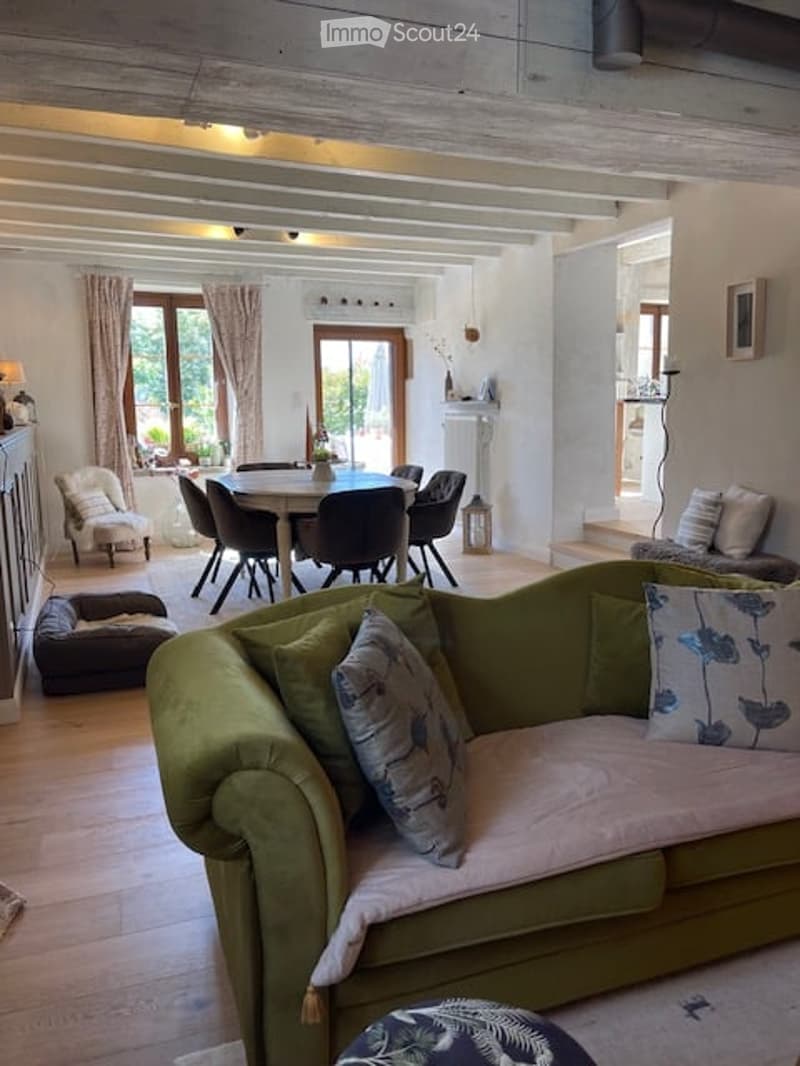 A cozy living room with a green sofa, a round dining table with chairs, a small dog bed, and a lamp on a side table. The room features wooden flooring and large windows with curtains.