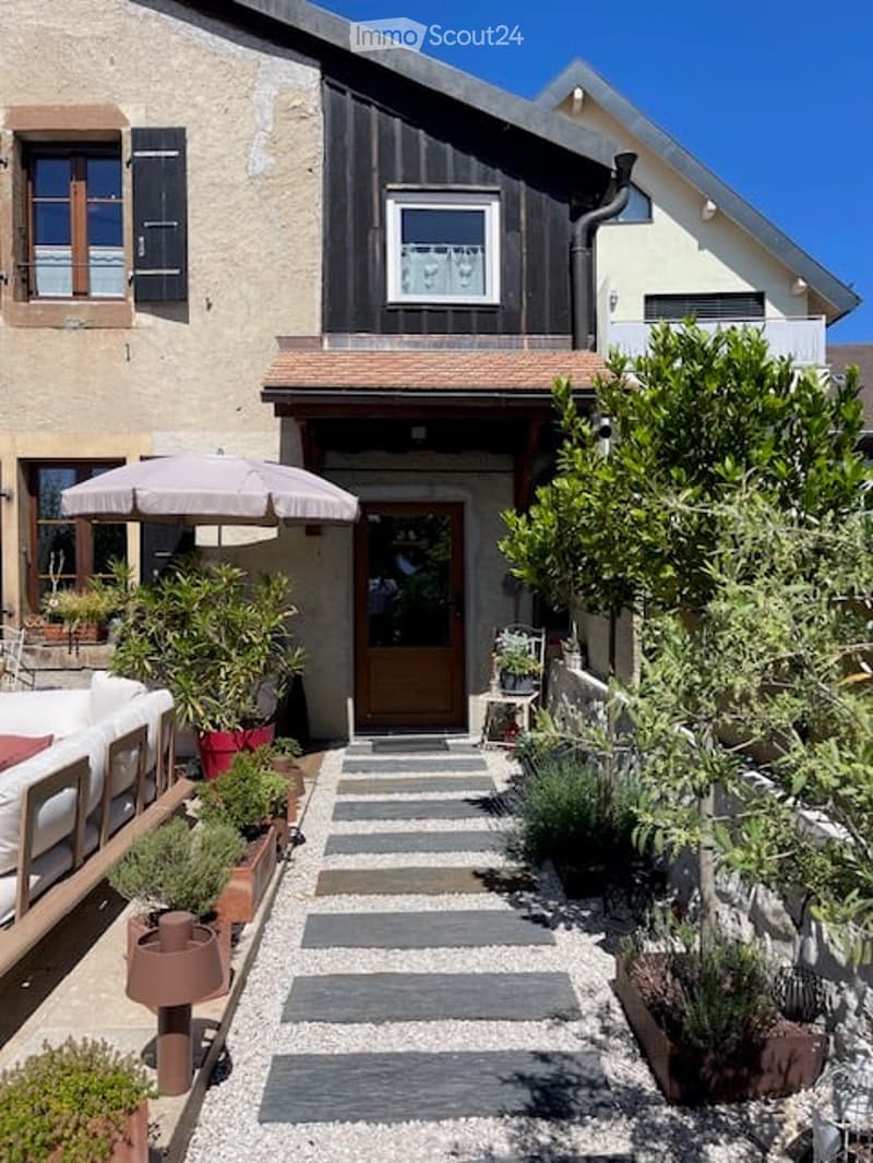 A charming garden path leads to the entrance of a house, flanked by potted plants and a small seating area with a white sofa and umbrella.