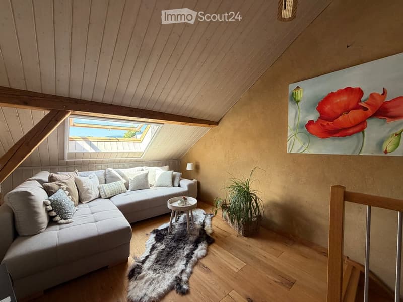 Cozy attic living room with wooden beams, skylight window, grey sectional sofa, small round table, potted plant, and large floral painting on the wall.
