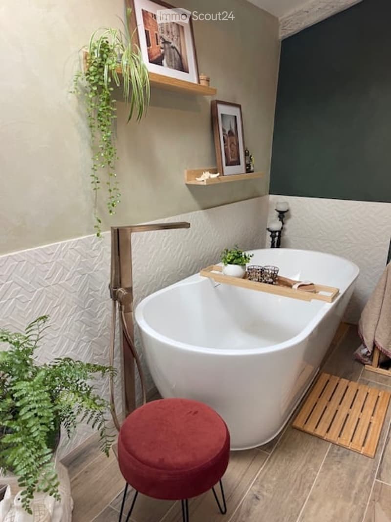 Modern bathroom with a freestanding bathtub, wooden tray on the edge, red velvet stool, shelves with framed pictures and plants, and a candle holder.