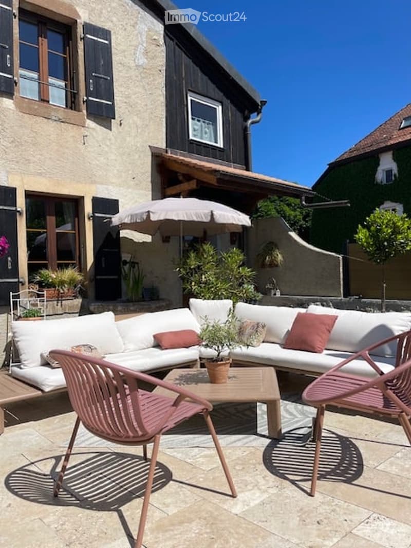 Outdoor patio with a white sectional sofa, pink cushions, a wooden coffee table, pink chairs, and a large umbrella. Potted plants and a stone house with shutters are visible in the background.