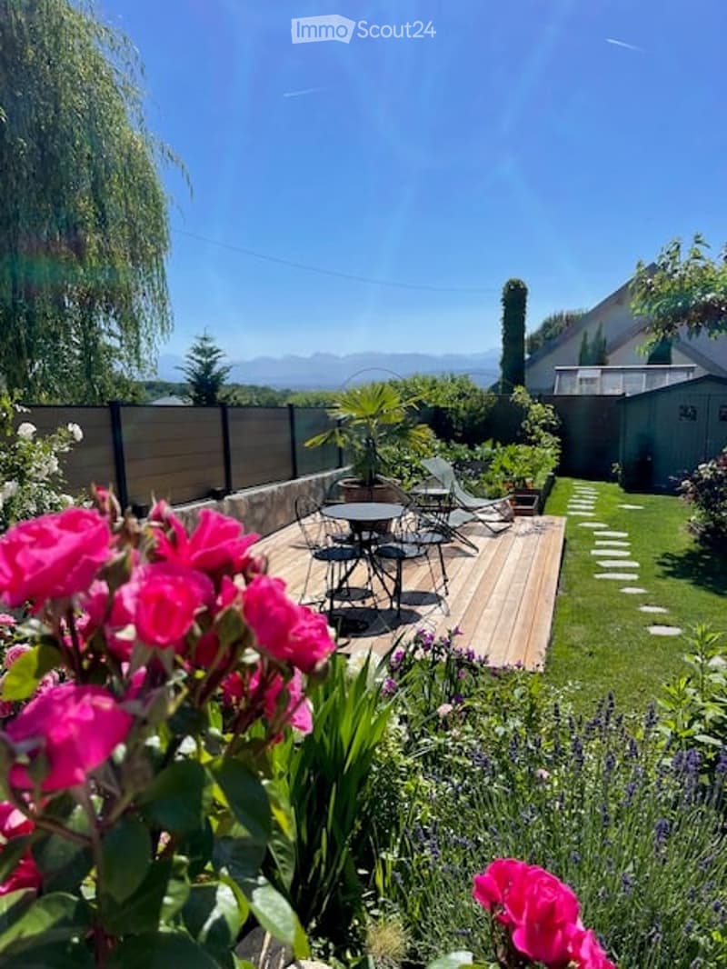 Well-maintained garden with a wooden deck, outdoor furniture, and colorful flowers, surrounded by greenery and a wooden fence.