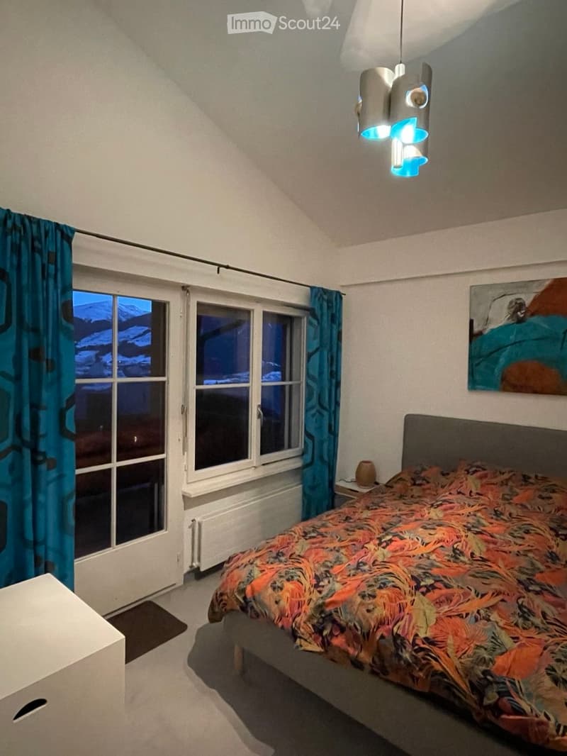 Cozy bedroom with a large bed, colorful bedspread, nightstand, painting on the wall, and blue curtains covering the windows.