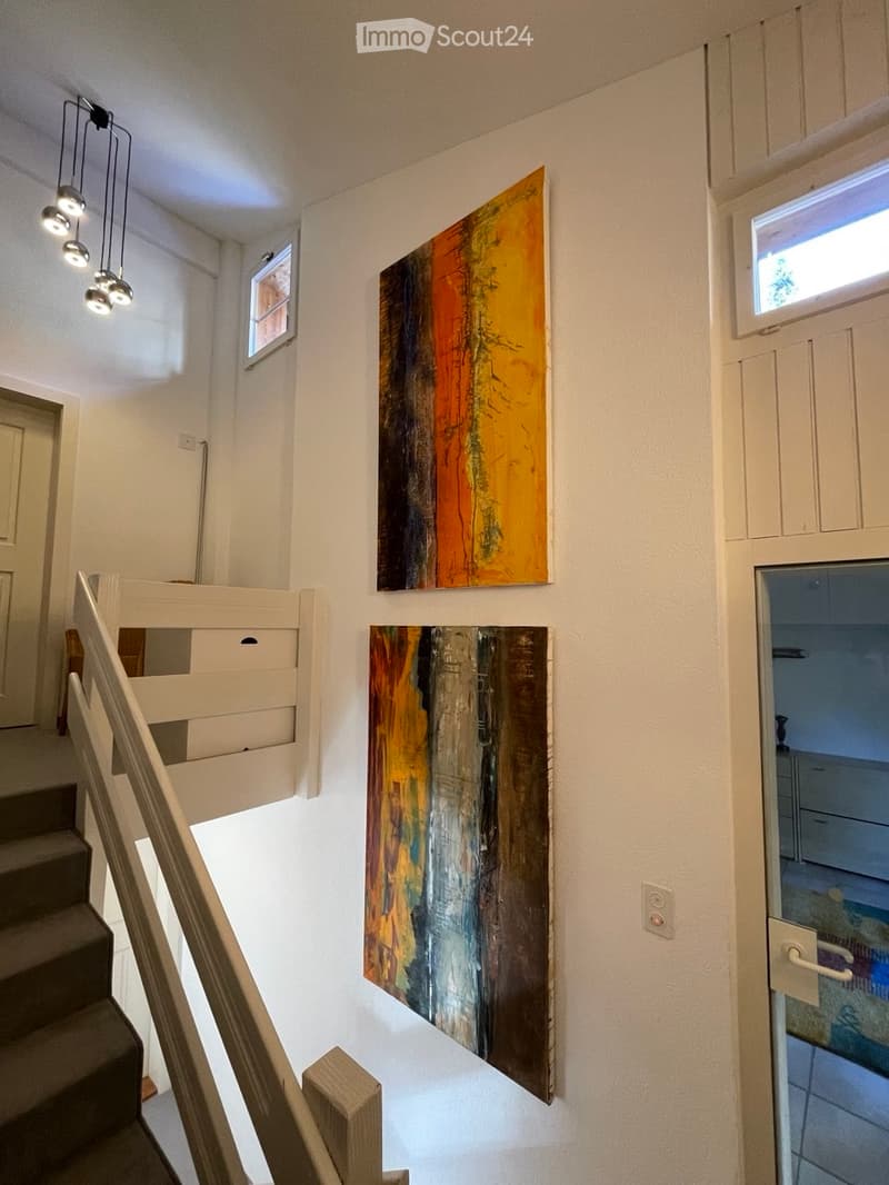 Staircase with modern wooden railing, colorful abstract paintings on walls, pendant lights, and a view into an adjacent room.