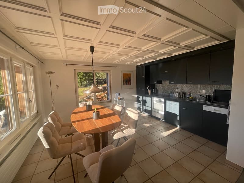 Bright, modern kitchen and dining area with dark cabinets, a wooden dining table, white chairs, and a pendant light. The kitchen features an oven, dishwasher, and sink.