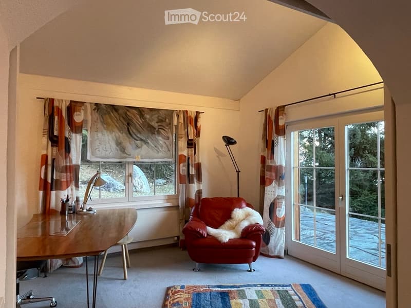 A cozy room with a slanted ceiling, featuring a red leather armchair with a white throw, a wooden table, a floor lamp, and large windows with colorful curtains.