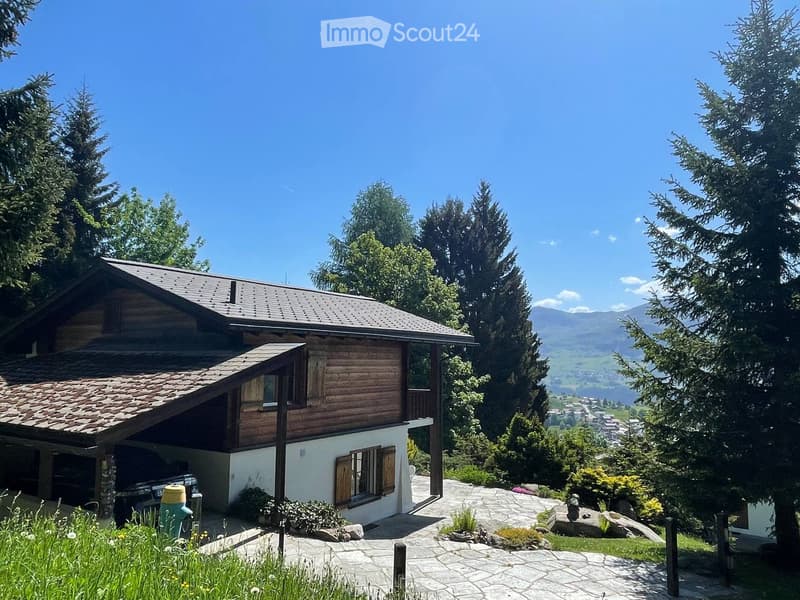 A wooden house with a tiled roof, surrounded by trees and a scenic mountain view in the background.