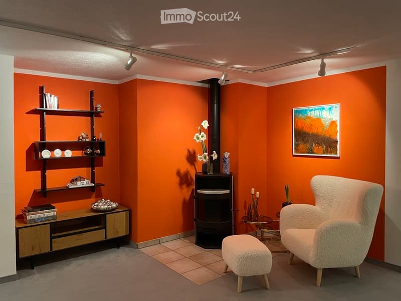 Modern living room with orange walls, a fireplace, a white armchair, a stool, a shelf with decorative items, and a small table with candles.