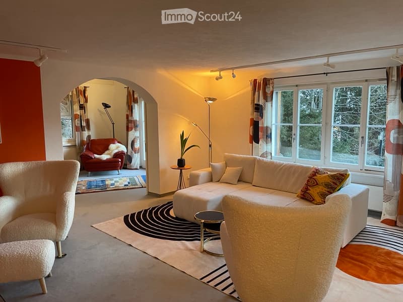 Modern living room with large windows, cream-colored sectional sofa, red leather armchair, cream-colored chair, coffee table, floor lamp, and potted plant.
