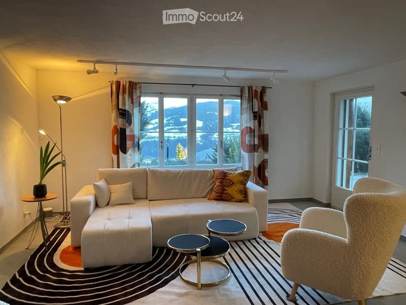 Modern living room with large windows offering a scenic view, featuring a white sectional sofa, decorative pillows, a round coffee table, a floor lamp, and a potted plant.