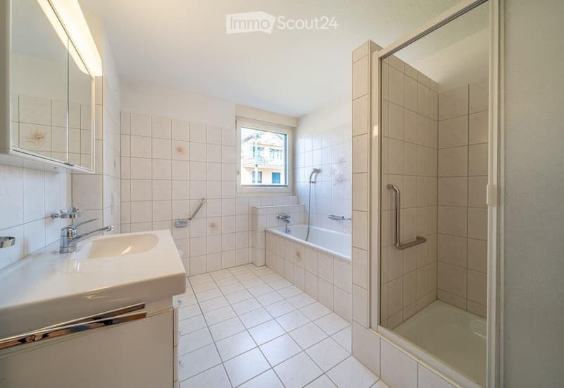 Bathroom. Bathroom with white tiled walls and floor, a sink with a modern faucet, a bathtub, a glass-enclosed shower stall, and a mirror above the sink. A window provides natural light.